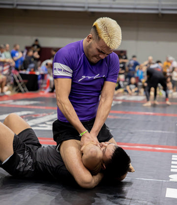 Two male grapplers compete in a Brazilian jiu-jitsu tournament on a black mat with a blurred audience