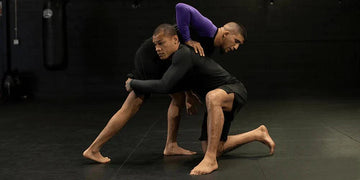 Two men in dark rash guards wrestling on a black mat in a gym with punching bags in the background