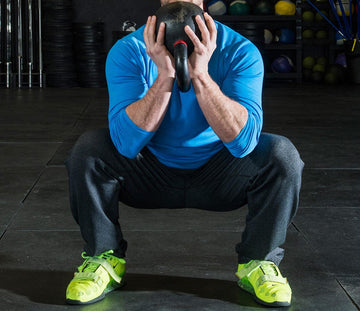 Man in blue shirt and gray pants doing kettlebell goblet squat in gym with fitness equipment background