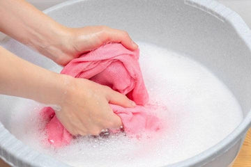 Hands hand washing a pink cloth in a basin filled with white soapy water