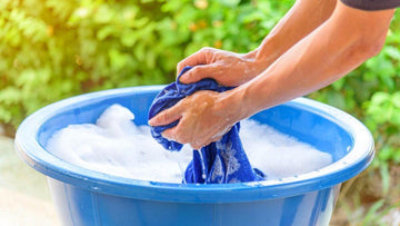 Hands washing blue fabric in a soapy water-filled blue plastic basin outdoors with green foliage background