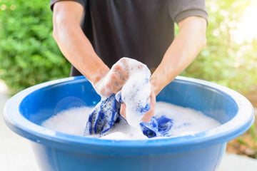 Hands washing blue fabric in a large blue basin filled with soapy water outdoors