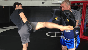 Two men training in MMA gym, one delivering a high kick while the other holds pads